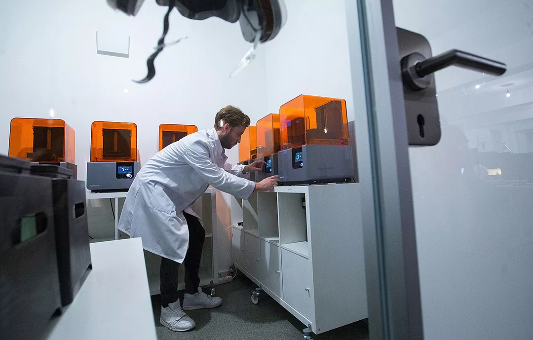 A WORKER IN A WHITE COAT BENDS DOWN TO OPERATE A MACHINE IN A ROOM FULL OF MACHINES