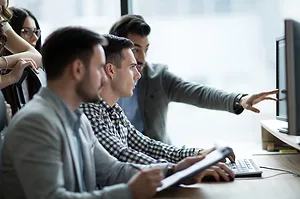 A TEAM OF PEOPLE SITTING AT A DESK ARE LOOKING AT A COMPUTER WITH ONE PERSON POINTING AT THE SCREEN. THE CLOSEST PERSON TO THE FRONT IS HOLDING A CLIPBOARD.