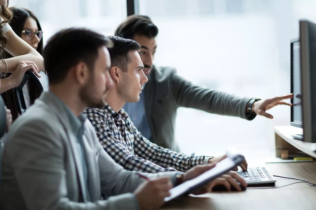 A TEAM OF PEOPLE SITTING AT A DESK ARE LOOKING AT A COMPUTER WITH ONE PERSON POINTING AT THE SCREEN. THE CLOSEST PERSON TO THE FRONT IS HOLDING A CLIPBOARD.