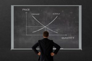 A MAN STANDS IN FRONT OF A BLACKBOARD WITH A CHART OF SUPPLY AND DEMAND 