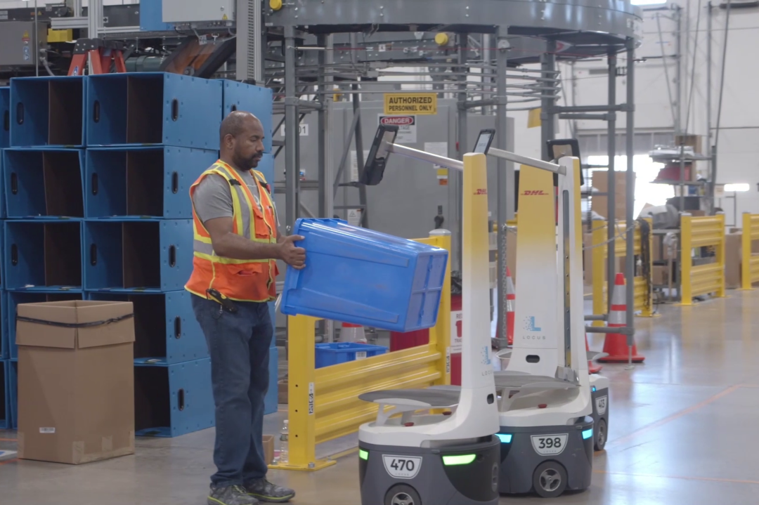 A WORKER LOADS A BIN ONTO A MOBILE ROBOT IN A DHL WAREHOUSE