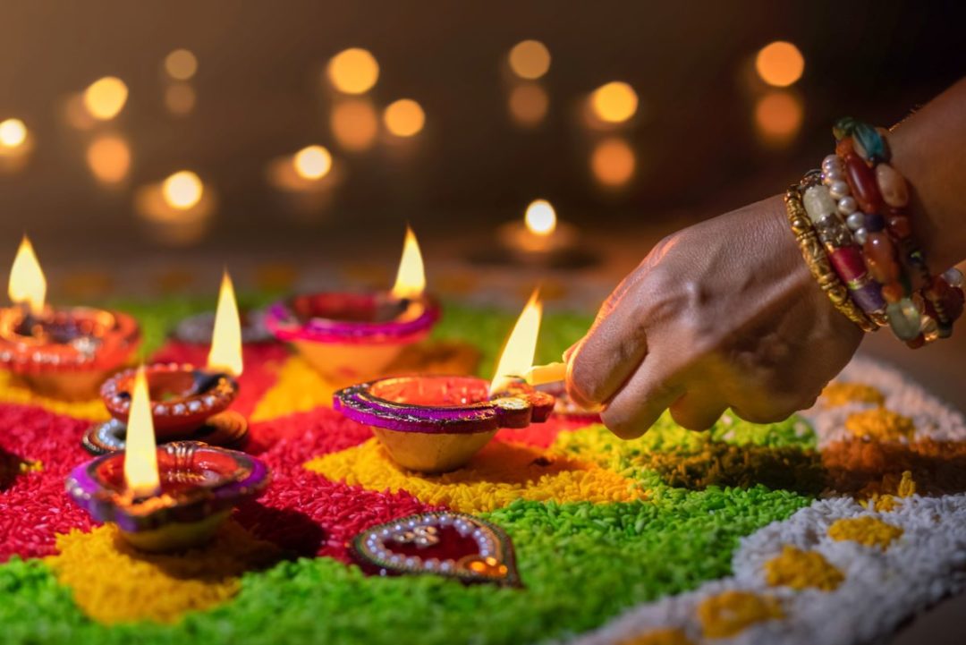 A BANGLED HAND LIGHTS ONE OF AN ARRAY OF BEJEWELLED DIWALI FESTIVAL LAMPS ON A BED OF COLORED SAND