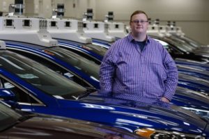 A MAN STANDS SMILING SURROUNDED BY SHINY NEW CARS