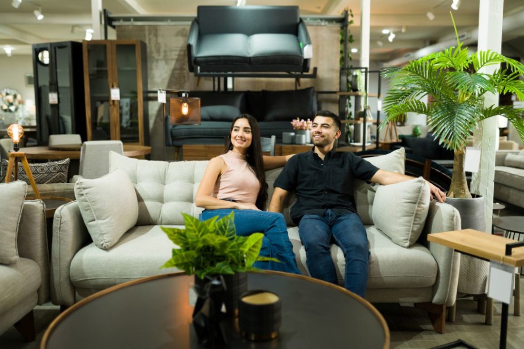 A COUPLE HOLD HANDS WHILE SITTING ON A SOFA SURROUNDED BY FURNITURE IN A STORE