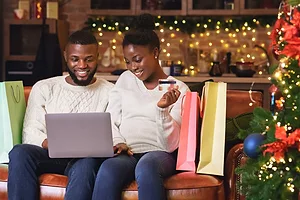 TWO PEOPLE SIT ON A SOFA NEXT TO A CHRISTMAS TREE SURROUNDED BY GIFT BAGS WHILE SMILING INTO AN OPENED LAPTOP COMPUTER
