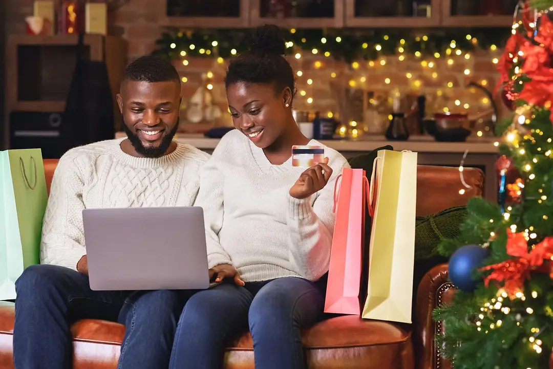 TWO PEOPLE SIT ON A SOFA NEXT TO A CHRISTMAS TREE SURROUNDED BY GIFT BAGS WHILE SMILING INTO AN OPENED LAPTOP COMPUTER