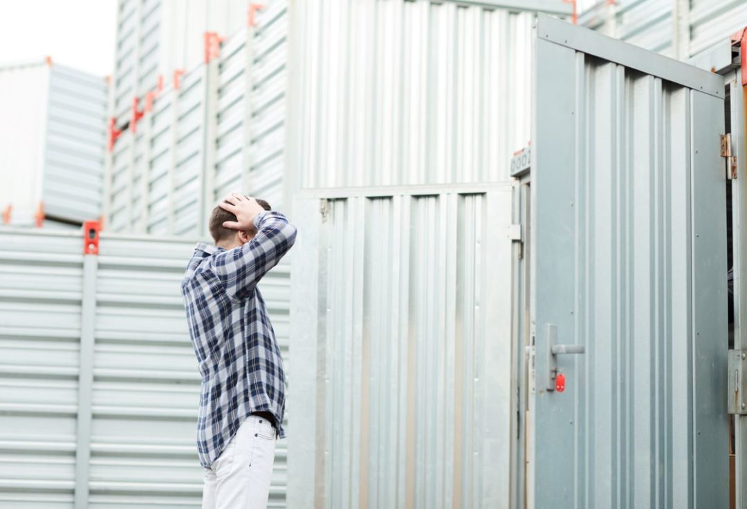 A MAN STANDS IN FRONT OF AN OPEN SHIPPING CONTAINER WITH HIS HANDS ON HIS HEAD IN FRUSTRATION