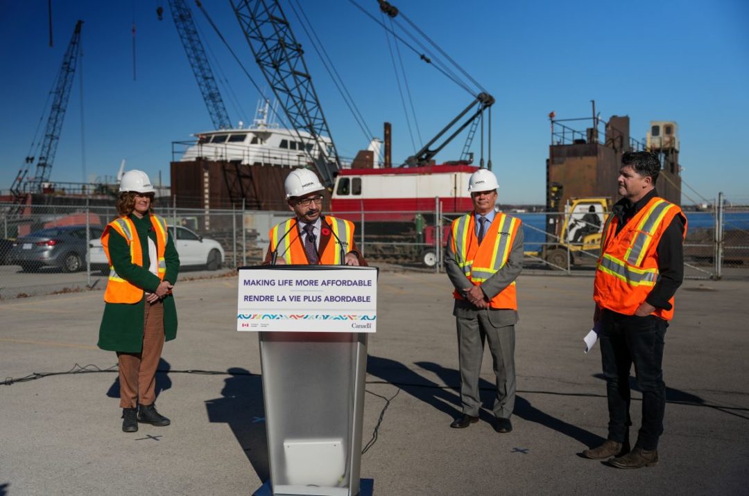 A PERSON STANDS AT A PODIUM SURROUNDED BY THREE OTHERS, ALL IN HI-VIS VESTS, AGAINST A BACKDROP OF PORT CRANES