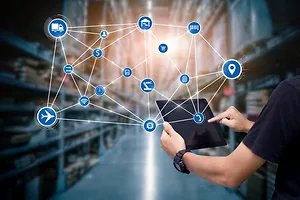 A PERSON HOLDING A TABLET COMPUTER CASTS AN IMAGINARY WEB OF CONNECTIONS ACROSS A WAREHOUSE AISLE