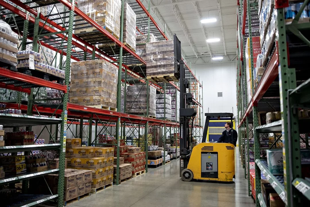 A WORKER OPERATES A FORKLIFT IN AN AISLE OF A WAREHOUSE