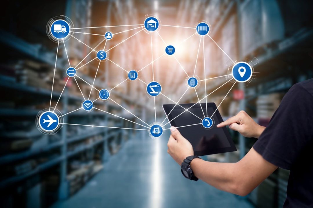 A PERSON HOLDS UP A TABLET COMPUTER IN A WAREHOUSE, SUPER-IMPOSED BY A GRAPHIC SHOWING A COMPLEX WEB OF SUPPLY CHAIN ELEMENTS