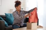 A WOMAN SEATED ON A COUCH PULLS A SWEATER OUT OF A DELIVERY BOX, FROWNING
