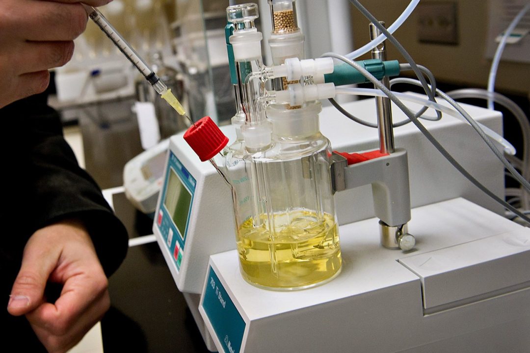 A HAND HOLDS A PIPETTE DEPOSITING YELLOW LIQUID INTO A BEAKER IN A LAB