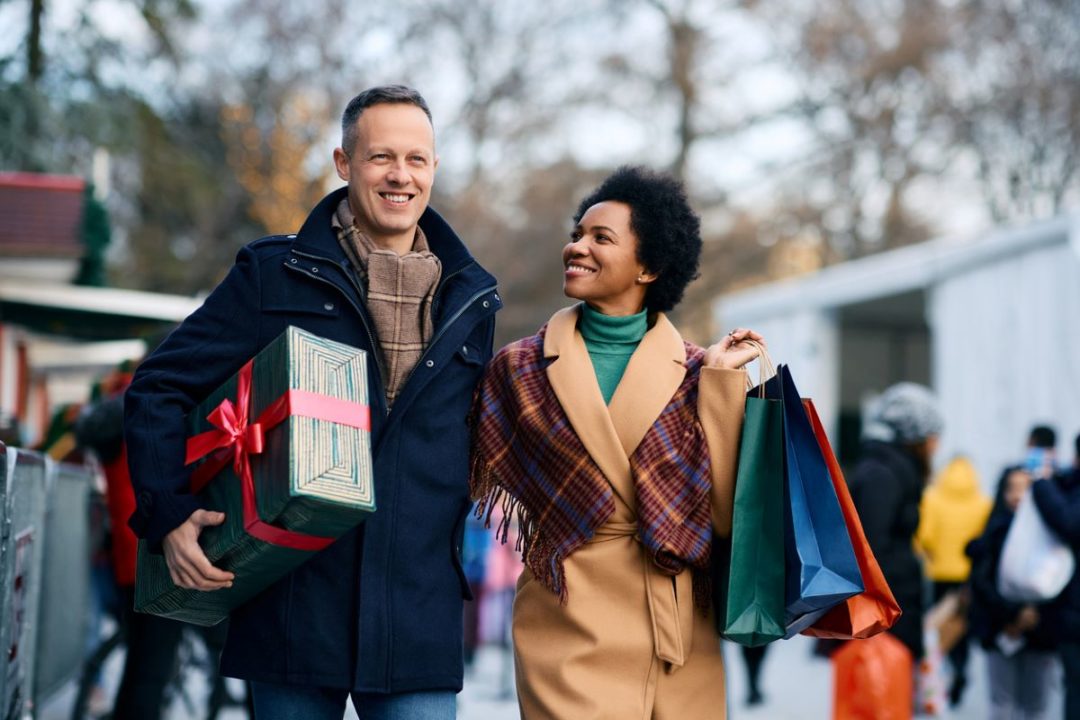 A MAN AND A WOMAN SMILE AS THEY WALK DOWN A STREET CARRYING BAGS OF SHOPPING