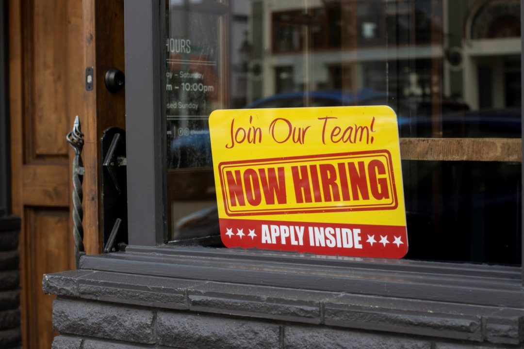 A SIGN SAYING "NOW HIRING" SITS IN A STORE WINDOW