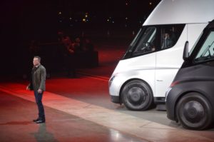 ELON MUSK STANDS ON A STAGE IN FRONT OF A TESLA SEMI TRUCK