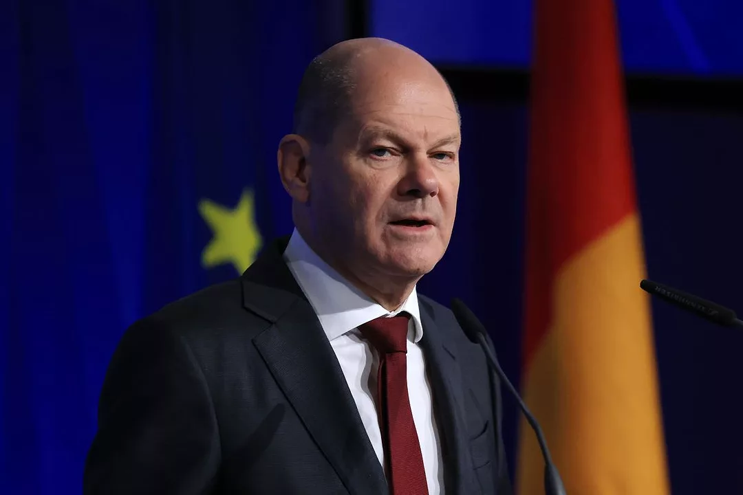 A MAN IN A JACKET AND TIE STANDS IN FRONT OF EU AND GERMAN FLAGS
