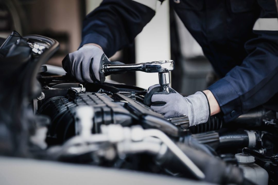 GLOVED HANDS WIELD A WRENCH ON AN ENGINE INSIDE THE HOOD OF A CAR