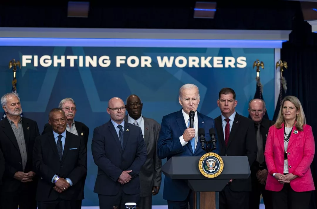 A LINE OF PEOPLE STAND BEHIND A PODIUM AND IN FRONT OF A SIGN READING "FIGHTING FOR WORKERS"
