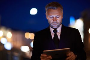 A MAN IN A SUIT STANDS, HIS FACE BATHED IN THE GLOW OF A TABLET COMPUTER, OUT OF FOCUS LIGHTS BEHIND HIM