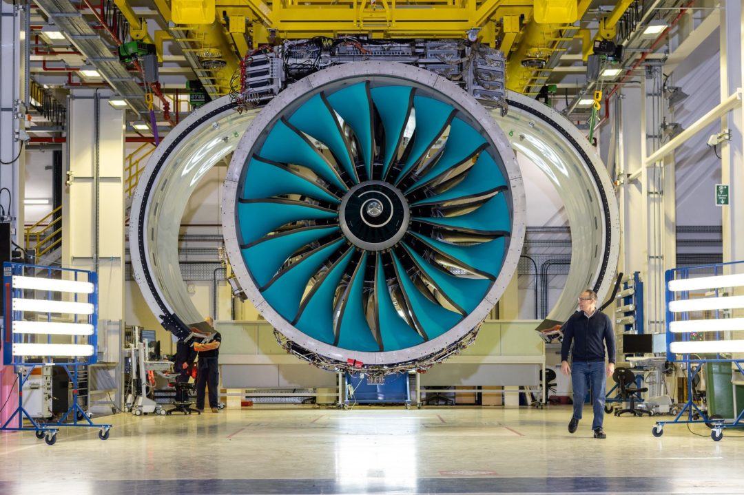 A HUGE JET ENGINE WITH BLUE FINS SITS IN A HANGAR, WITH STAFF MILLING ABOUT ON THE GROUND