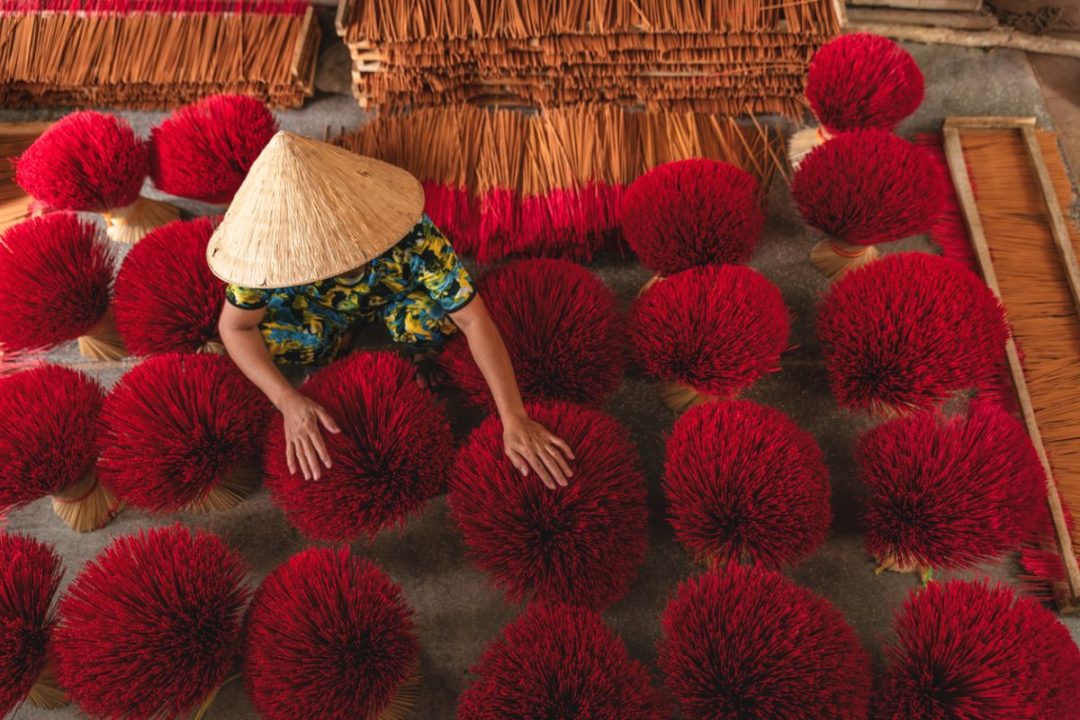 A WOMAN IN A CONICAL STRAW HAT TENDS BRIGHT RED CLUSTERS OF INCENSE