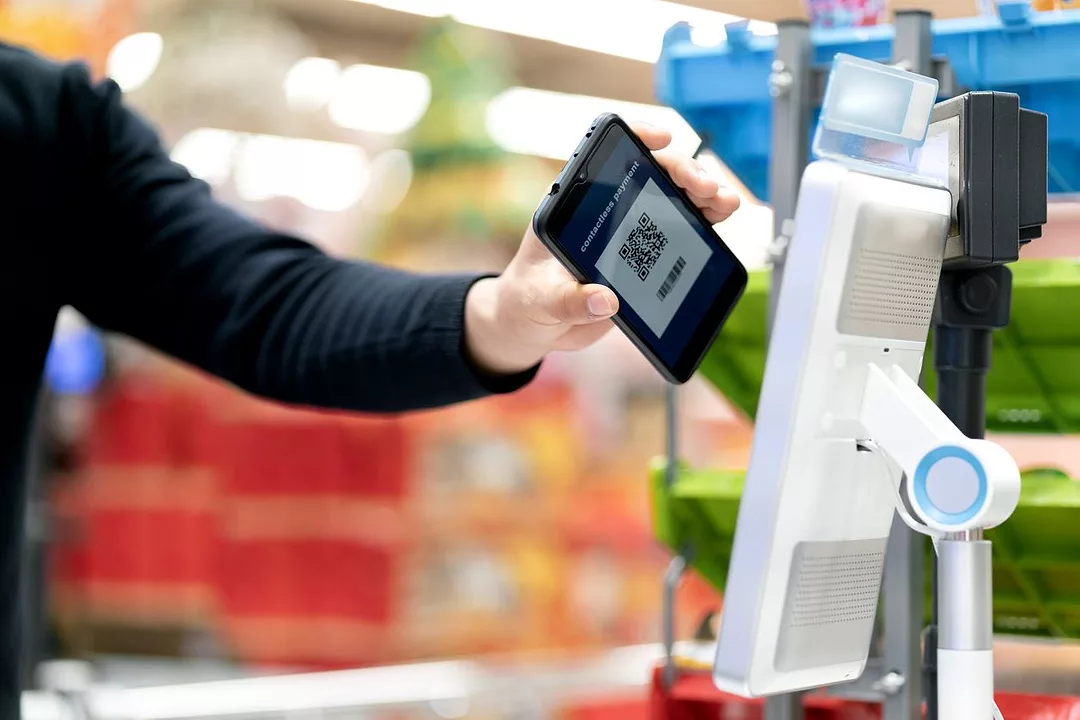 A SHOPPER HOLDS UP A PHONE WITH A QR CODE TO A READER AT A SUPERMARKET CHECKOUT