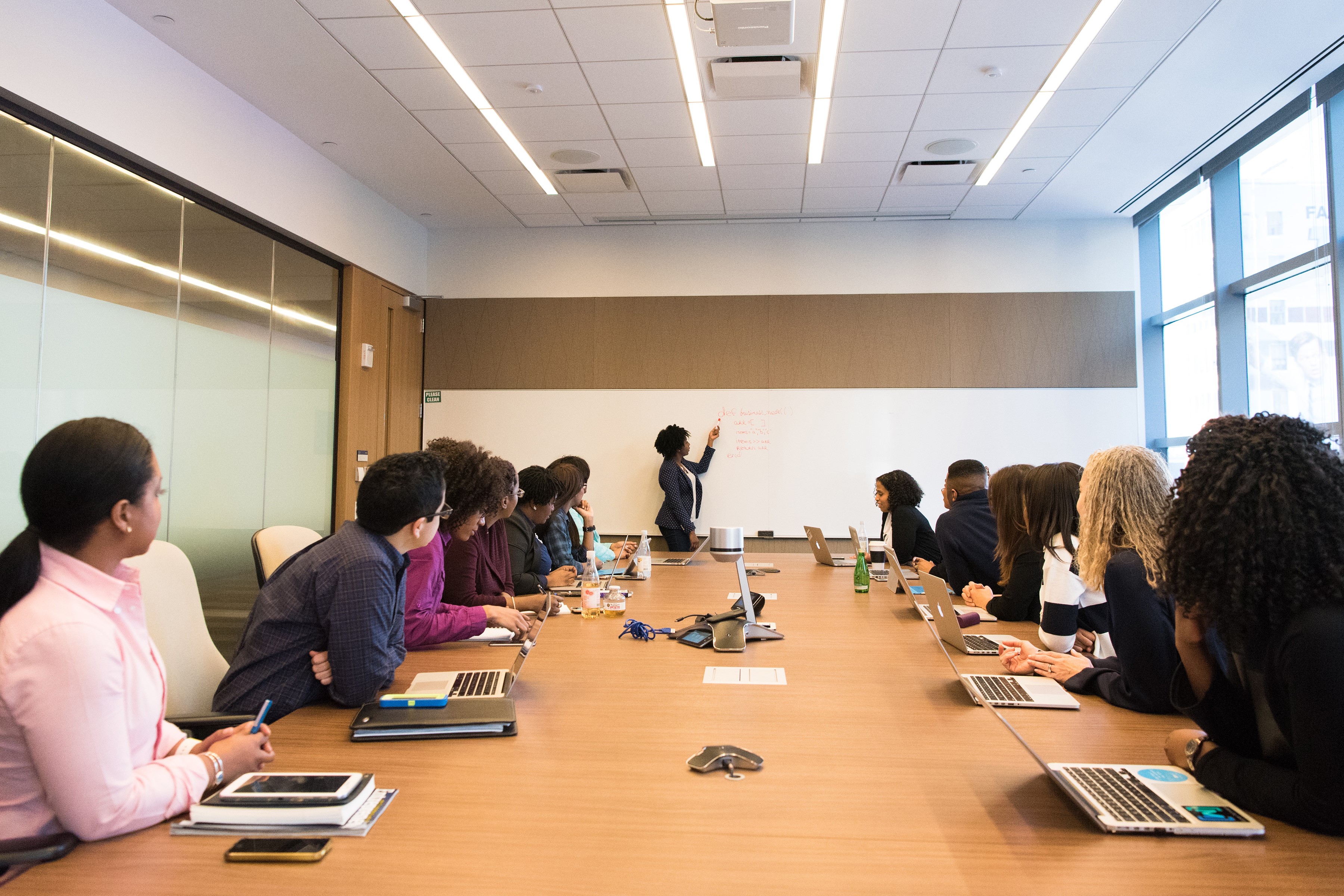 A WOMAN LEADS A MEETING AT THE HEAD OF A LONG TABLE OF OFFICE WORKERS, DRAWING ON A WHITE BOARD
