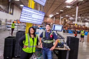 TWO SMILING WORKERS IN HI-VIS VESTS STAND SIDE BY SIDE IN A GIANT WAREHOUSE