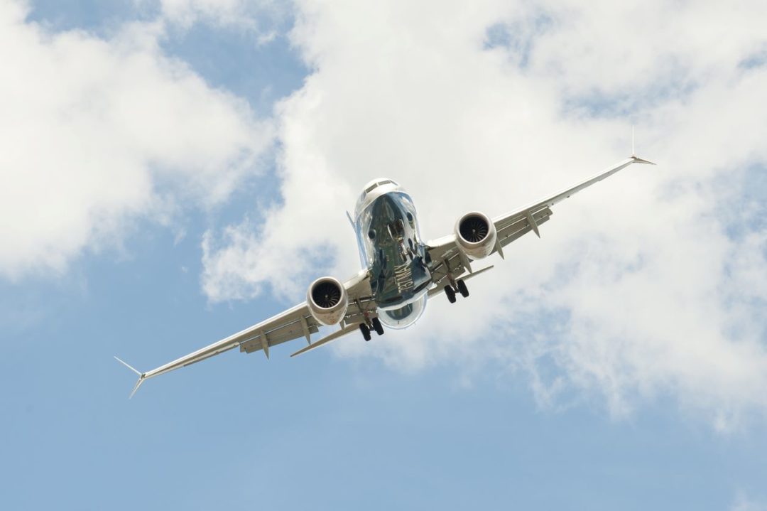 A BOEING 737 MAX, SEEN FROM BELOW, FLIES THROUGH A BLUE SKY WITH CLOUDS