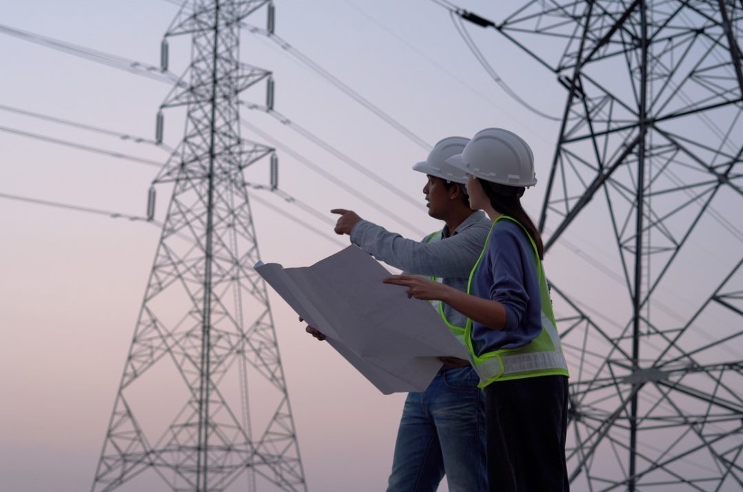TWO WORKERS DISCUSS A DOCUMENT UNDER TOWERING ELECTRICITY PYLONS