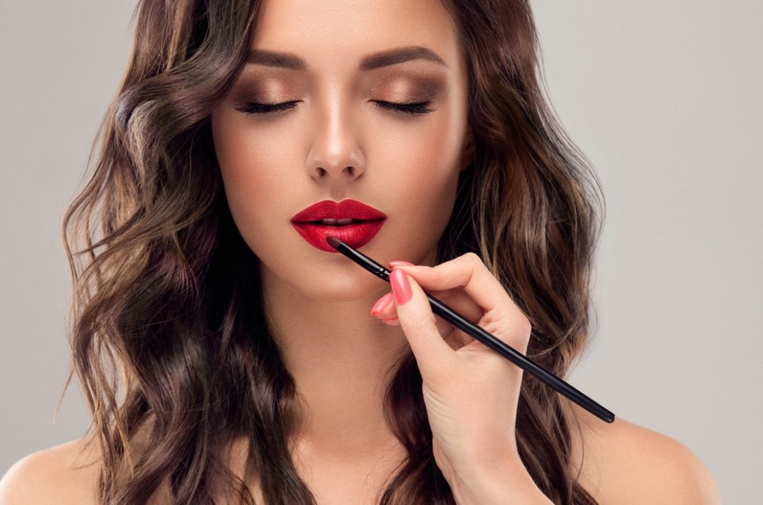 AN ELEGANT HAND APPLIES LIPSTICK WITH A BRUSH TO A GLAMOROUS WOMAN'S FACE