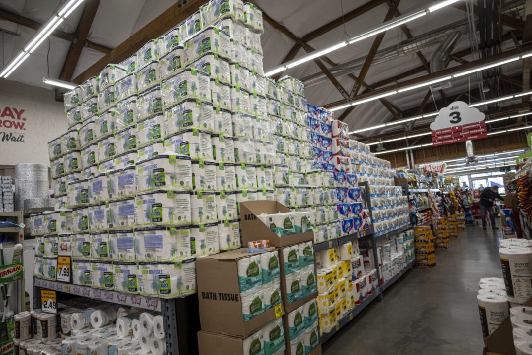 A GIANT PYRAMID OF VARYING BRANDS OF TOILET PAPER SITS AT THE END OF A DISCOUNT STORE AISLE