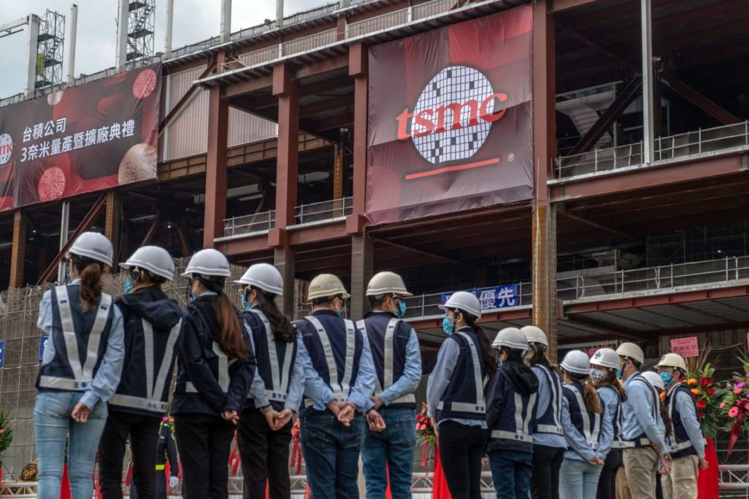 WORKERS IN HARDHATS STAND IN A LINE UNDER A TSMC SIGN