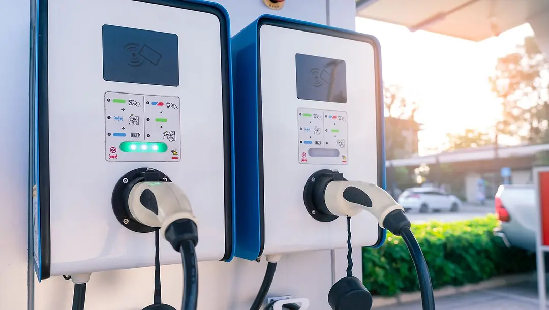 TWO EV CHARGING STATIONS SIT SIDE BY SIDE AT A GAS STATION, A CAR OUT OF FOCUS IN THE DISTANCE
