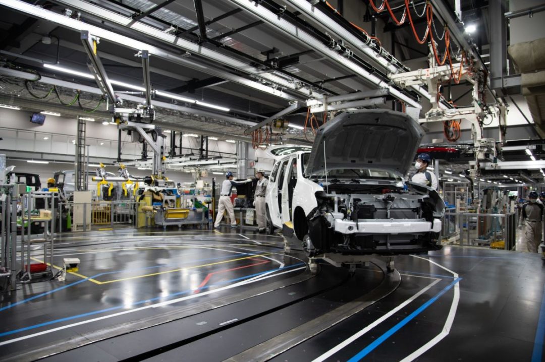 WORKERS ADVANCE A PARTIALLY MANUFACTURED CAR THROUGH AN ASSEMBLY LINE