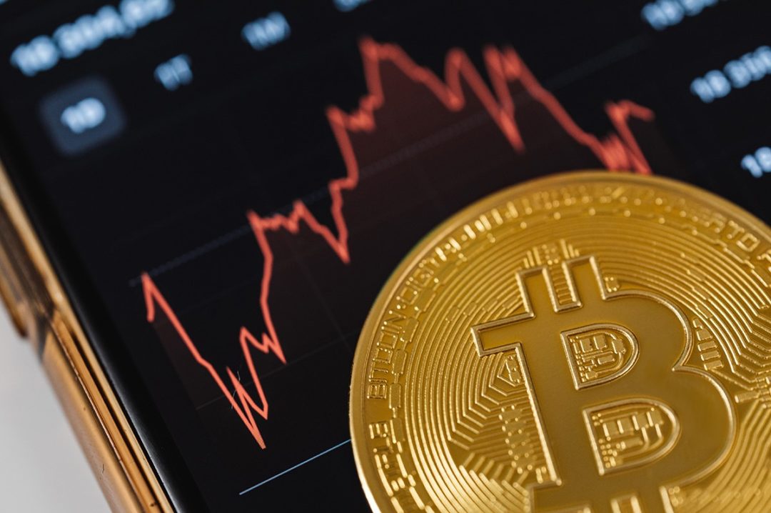 A BITCOIN SYMBOL SITS BELOW A GRAPH SHOWING WILD FLUCTUATIONS
