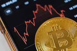 A BITCOIN SYMBOL SITS BELOW A GRAPH SHOWING WILD FLUCTUATIONS