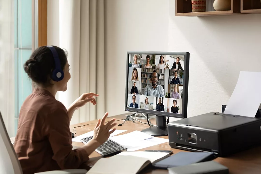 A WOMAN IN A HOME OFFICE SITS IN FRONT OF A COMPUTER SCREEN WITH MULTIPLE IMAGES OF PEOPLE IN CONFERENCE