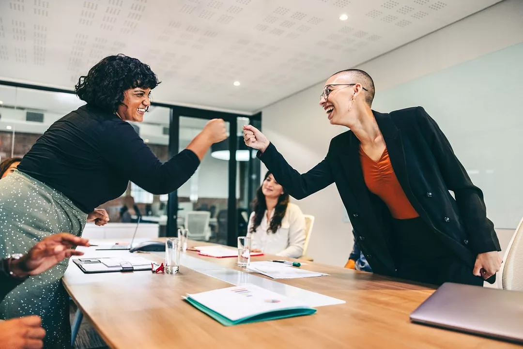TWO WOMEN FIST-BUMP OVER A TABLE IN A CONFERENCE ROOM