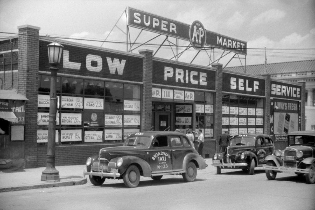 A BLACK AND WHITE PHOTO SHOWS 40S ERA CARS PARKED IN FRONT OF AN A&P SUPERMARKET DISPLAYING A SIGN THAT SAYS LOW PRICE SELF SERVICE