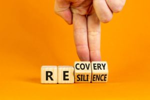 TWO FINGERS MANIPULATE WOODEN LETTER BLOCKS TO TURN FROM SHOWING THE WORD RECOVERY TO RESILIENCE