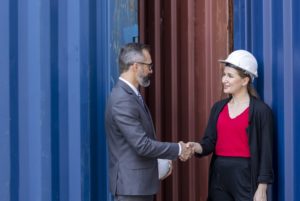 A MAN IN A SUIT SHAKES HANDS WITH A WOMAN IN A HARD HAT, NEXT TO A STACK OF CONTAINERS