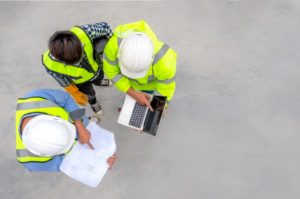 THREE CONSTRUCTION WORKERS IN HARD HATS, SEEN FROM ABOVE, CONSULT BLUEPRINTS