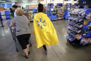 TWO PEOPLE PUSH A SHOPPING CART THROUGH A STORE, ONE WEARING A CAPE BEARING THE WALMART LOGO