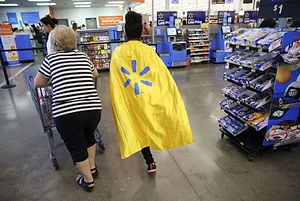 TWO PEOPLE PUSH A SHOPPING CART THROUGH A STORE, ONE WEARING A CAPE BEARING THE WALMART LOGO