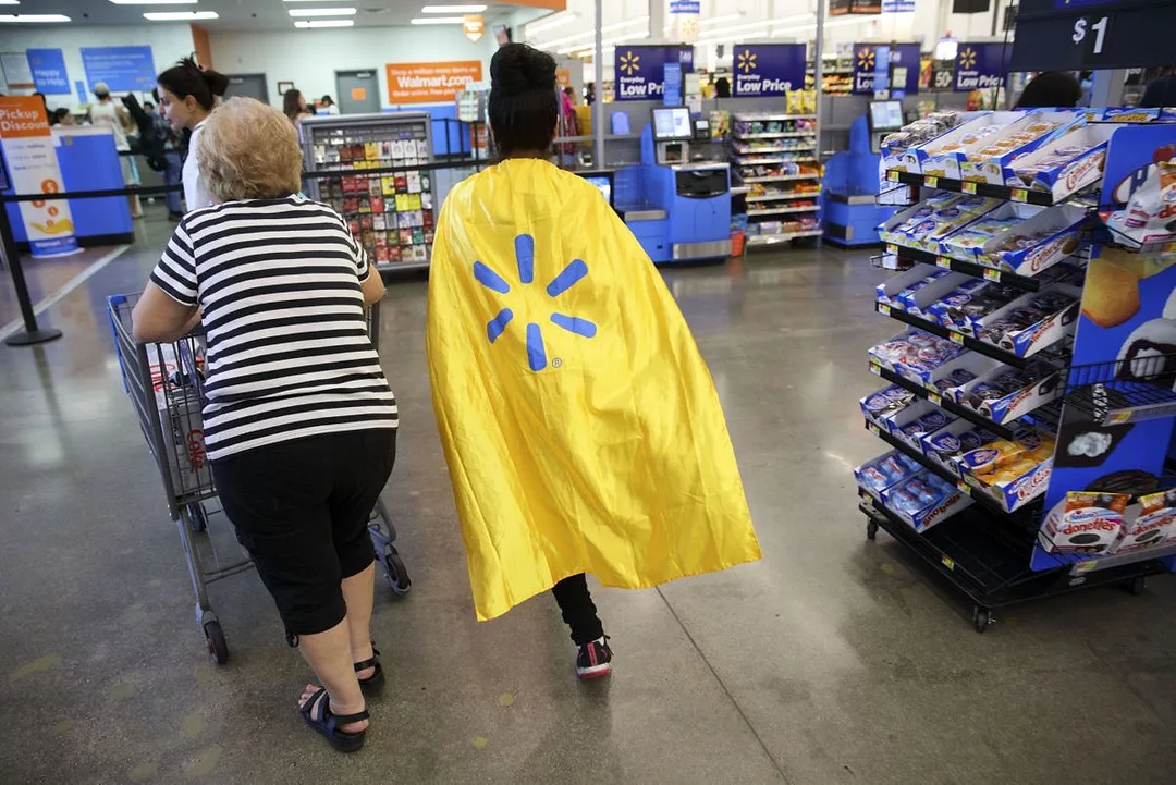TWO PEOPLE PUSH A SHOPPING CART THROUGH A STORE, ONE WEARING A CAPE BEARING THE WALMART LOGO