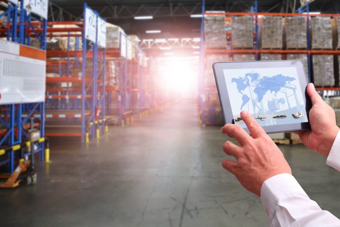 INSIDE A WAREHOUSE, TWO HANDS HOLD A TABLET COMPUTER SHOWING A MAP OF THE WORLD