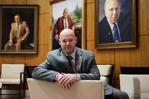 A MAN IN A SUIT SITS LEANING OVER THE BACK OF A CHAIR IN A WOOD-PANELLED ROOM FULL OF LARGE PHOTOS OF OTHER MEN IN SUITS