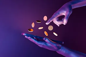 A GRAPHICALLY ALTERED IMAGE, SHOWING A PAIR OF HANDS HOLDS A DEVICE, SHOWERING IT WITH COINS